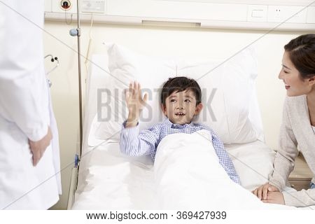 Happy Five Year Old Asian Kid Lying In Bed In Hospital Ward Waving Good-bye To Doctor