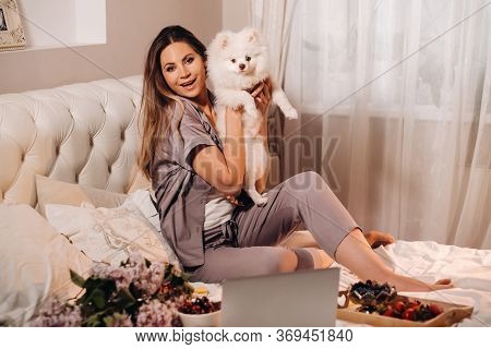 A Girl In Pajamas Sits In Bed At Night With Her White Dog Watching A Laptop And Eating Sweets.girl W