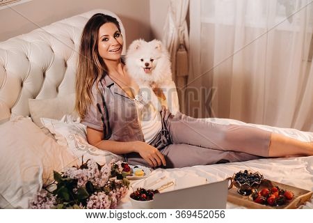 A Girl In Pajamas Sits In Bed At Night With Her White Dog Watching A Laptop And Eating Sweets.girl W
