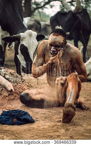 Mundari Tribe, South Sudan - March 11, 2020: Man Sitting On Ground Near Bonfire And Traditionally Sm