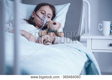 Girl Suffering From Pneumonia Lying In A Hospital Bed With Oxygen Mask And Teddy Bear