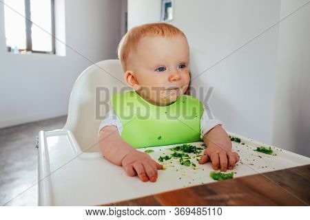 Portrait Of Dirty Baby Girl Sitting On Highchair At Dining Table. Little Child Starting Eating By He