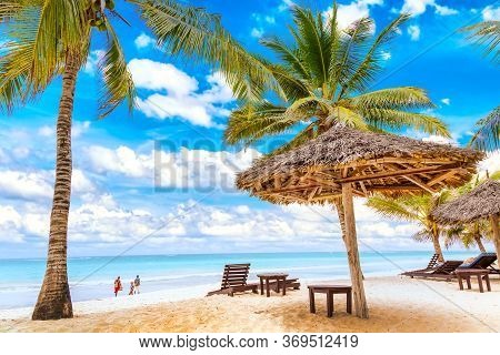 Sun Loungers Under Umbrella And Palms On The Sandy Beach By The Ocean And Cloudy Sky. Vacation Backg