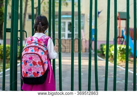 Little School Girl Sitting Next To School Fence Waiting For Going Back To Clases After Pandemic Outb