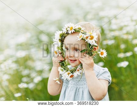 A Baby In A Light Blue Dress Looks Away Through A Wreath Of Cammomile. Stands In A Chamomile Field