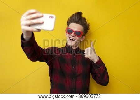 Guy Takes Selfie On Smartphone And Shows Like On Yellow Isolated Background, Hipster In Glasses Take
