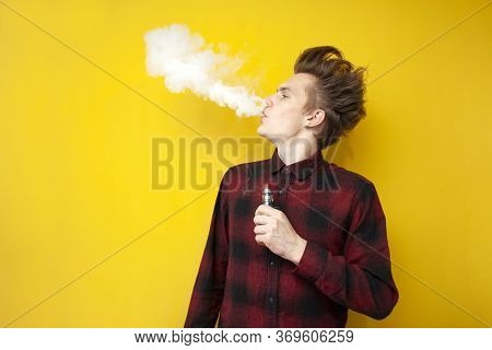 Guy Hipster Smokes An Electronic Cigarette On A Yellow Isolated Background, A Man Exhales A Cloud Of