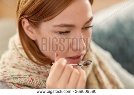 people, health and cold concept - sad sick woman in blanket and scarf taking cough syrup or antipyretic medicine at home