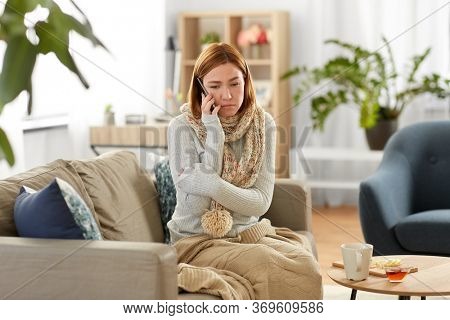 people, health and cold concept - sad sick woman in blanket and scarf calling on smartphone at home