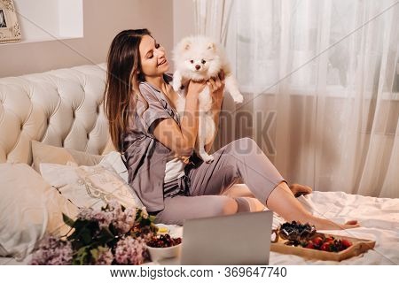 A Girl In Pajamas Sits In Bed At Night With Her White Dog Watching A Laptop And Eating Sweets.girl W