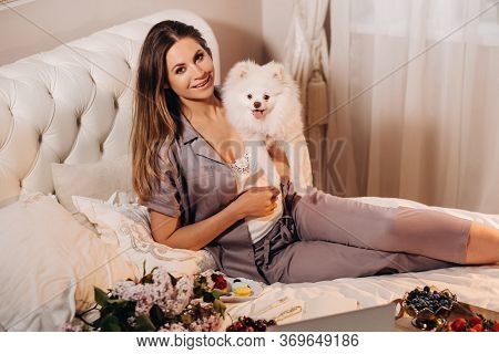 A Girl In Pajamas Sits In Bed At Night With Her White Dog Watching A Laptop And Eating Sweets.girl W