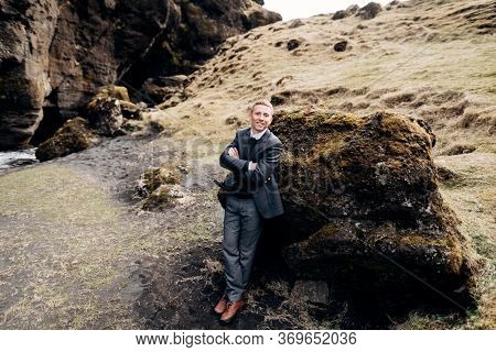 Portrait Of A Groom Leaning On A Stone In A Moss, Next To A Rocky Cleft With A Mountain River. Desti