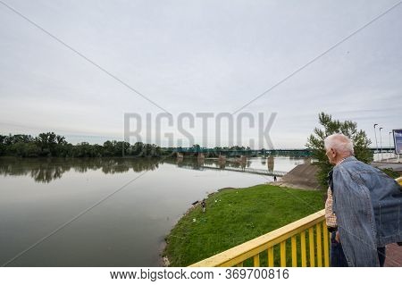 Brcko, Bosnia - May 6, 2017: Old Senior Man Looking At A Steel Bridge Crossing The Sava River At The