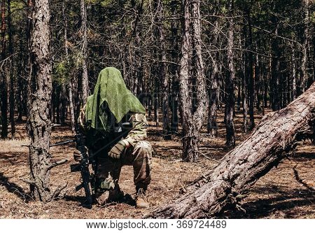 Photo of a fully equipped camouflaged forest sniper with rifle and tactical net scarf sitting in woods.
