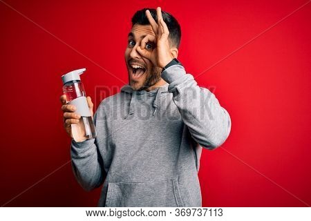 Young handsome sportsman drinking bottle of water to refeshment over red background with happy face smiling doing ok sign with hand on eye looking through fingers
