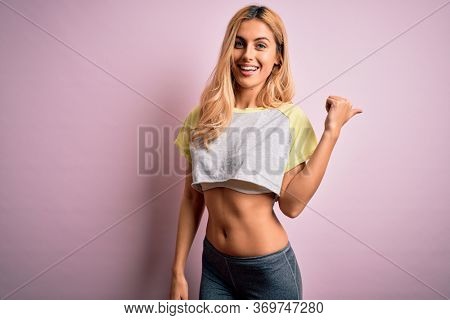 Young beautiful blonde sportswoman doing sport wearing sportswear over pink background smiling with happy face looking and pointing to the side with thumb up.