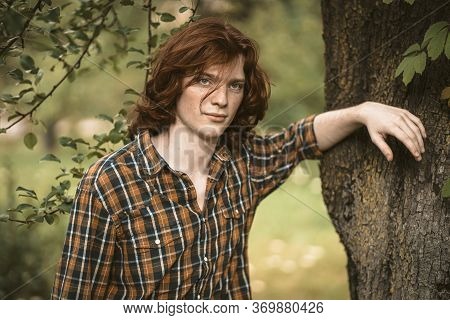 Foxy Man With Long Carroty Hair Stands Leaning On A Tree Trunk. Young Handsome Freckled Guy In Chequ