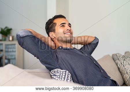 Young indian man at home relaxing and thinking with hands behind head. Middle eastern pensive guy resting at home. Latin thoughtful man sitting on couch while looking up.