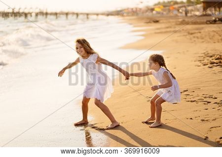 Girl Children In White Dresses Playing On The Beach. Older Sister Pulls The Younger Into The Water. 