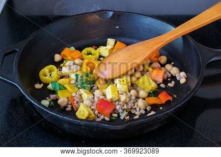 Stirring Bell Peppers, Tofu, Farro And Garbanzo Beans Mix In Cast Iron Skillet With A Wooden Spoon- 