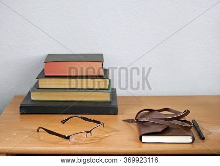 Leather Journal, Pencil, Readers, And Hardbound, Used Books Stacked On A Weathered, Used Wooden Desk