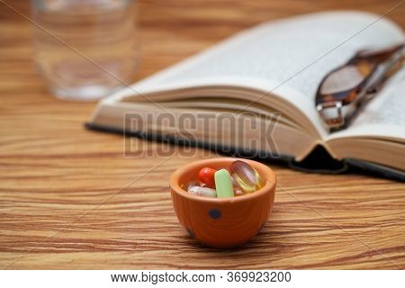 An Old Hardbound Book, Pair Of Broken Reader Glasses On It, A Bunch Of Pills In A Container, On Wood