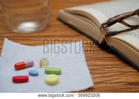 An Old Hardbound Book, Pair Of Broken Reader Glasses On It, A Bunch Of Pills On White Paper Towel, O