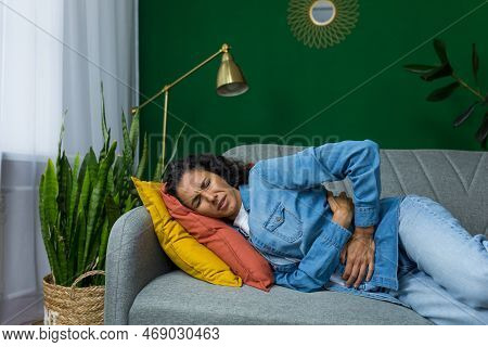 Hispanic Woman Sick Lying On Sofa In Living Room, Woman Has Severe Stomach Pain, Upset Holding Hands