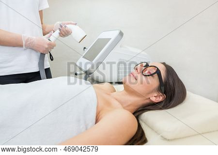 Women's Beauty. The Beautician Is Holding Aparatlaser Epilation Near A Beautiful Girl In A Medical C