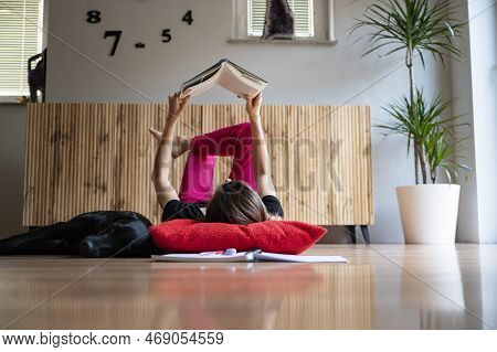 Low Angle View Of A Young Woman, A Student, Lying On A Living Room Floor With Her Head On Red Pillow