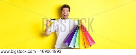 Happy Attractive Man Holding Shopping Bags, Showing Credit Card, Standing Over Yellow Background