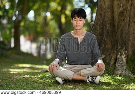 Pleasant Asian Man Sitting In Lotus Pose Sitting On Green Grass. Meditation And Mental Health Concep
