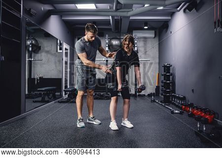 Fit Woman In Electric Muscular Suit To Stimulate Exercise At Training Center. A Male Trainer Helps H