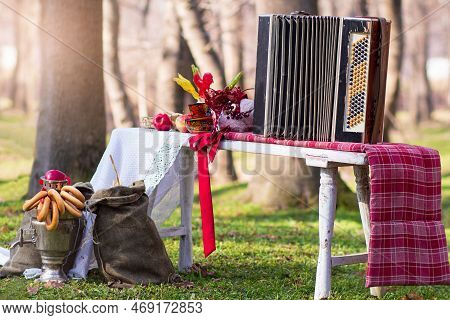 Samovar With Bagel, Candy, Accordion On White Tablecloth. Sack On Grass In Forest. Shrovetide, Folk 