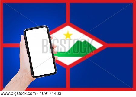Close-up Of Male Hand Holding Smartphone With Blank On Screen, On Background Of Blurred Flag Of Sint
