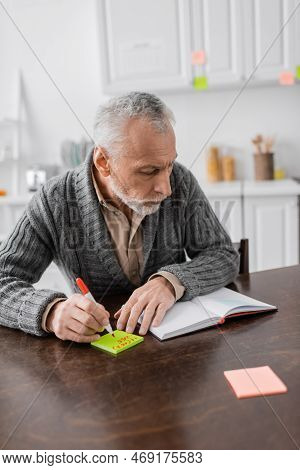 Grey Haired Man With Azheimers Syndrome Writing Phone Number On Sticky Notes Near Blank Notebook On 