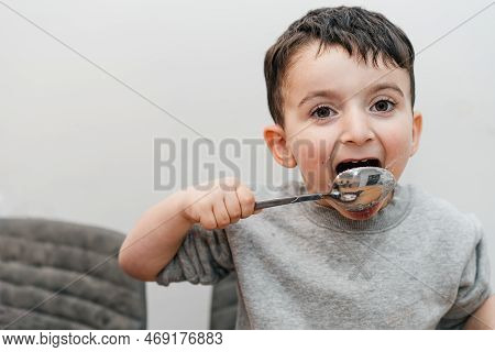 Funny Kid Licks A Spoon With Sugar. Portrait Joyful Beautiful Boy Eating Sugar And Looking At Camera