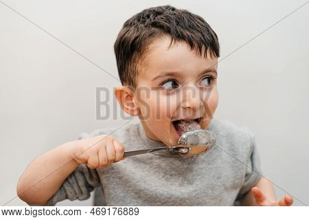 Funny Child Licks A Spoon With Sugar. Portrait Joyful Beautiful Boy Eating Sugar And Looking Away.