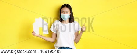 Covid-19, Pandemic And Real Estate Concept. Pretty Asian Woman Smiling In Medical Mask, Showing Pape