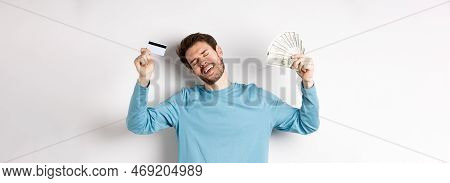 Handsome Man Dancing With Money And Plastic Credit Card, Standing In Casual Clothes Over White Backg