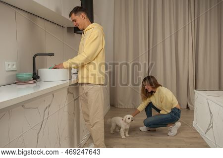 Happy Cherrful Couple In The Kitchen In Yellow Hoodies, Man Washing The Dishes And Woman Sitting And