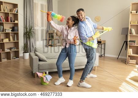 Happy Joyful Black Husband And Wife Tidying Home And Singing During Housecleaning, Using Mop And Dus