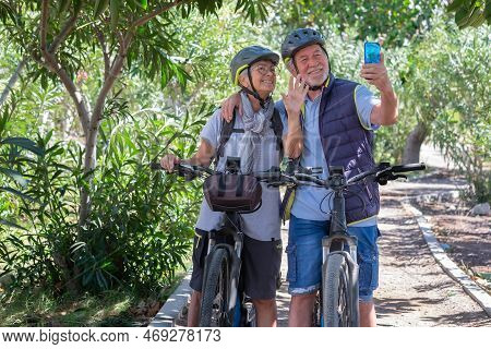 Active Caucasian Senior Couple With Electrobikes Standing Outdoors In The Park Video Calling By Cell