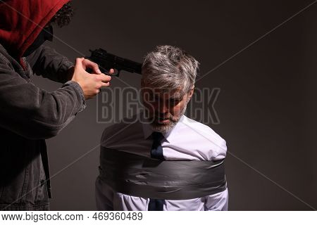 Kidnapper Aiming Gun At Tied Victim On Dark Background. Hostage Taking
