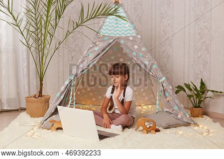 Portrait Of Joyful Dark Haired Little Girl With Pigtails Lying In Peetee Tent With Laptop, Having Vi