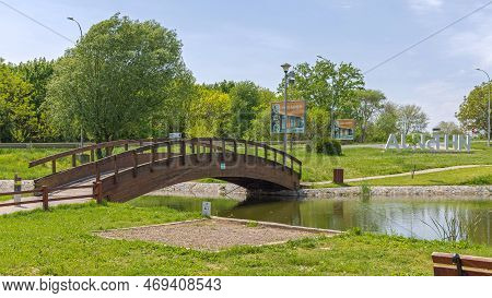 Indjija, Serbia - May 08, 2022: Wooden Arch Bridge Over Fishing Pond For Pedestrians In Town Park.