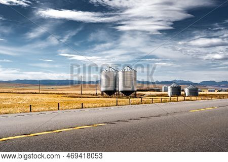 Pair Of Steel Grain Silos Standing Next To A Divided Highway Along Harvested Fields And Distant Rock