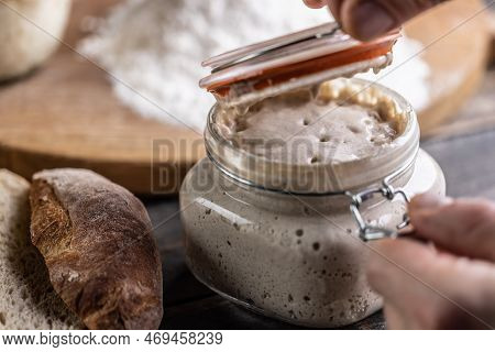 Male Hands Open A Jar With Active Yeast, Flour And Fresh Bread And Pastries In The Background