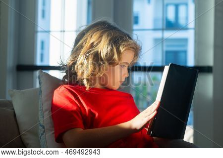 Child Reading Book At Home. Little Boy Sitting On Couch In Living Room Reading Story Book. Kid Doing