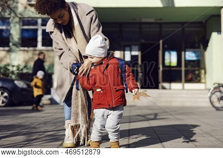 Babysitter Putting Backpack On Kindergarten Child In The Schoolyard. Babysitter Standing In The Scho